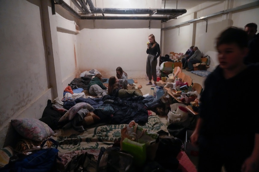 People lie and sit on mattresses on the floor of a dimly-lit basement, while some others stand.