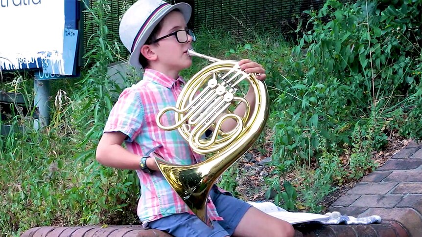 A young French Horn player sits on a brick wall in front of some greenery and plays french horn.