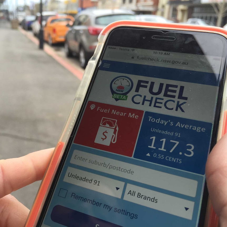 A smart phone held by a person on the roadside. The phone display shows the Fuel Check website