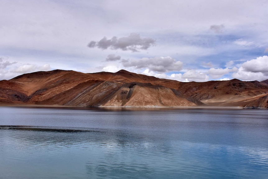 A pristine blue lake with bare, jagged hills behind on a cloudy day.