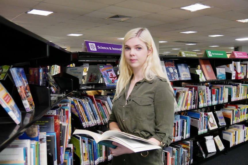Tutor Angela Fleming holds a book and stands in front of a library shelf.