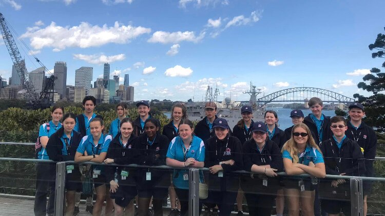 A group of students wearing turquoise and blue t-shirts stand in front of Sydney Harbour Bridge, Sydney skyline.