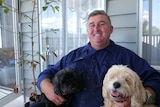 A man sits on a couch with two dogs on either side of him and smiles