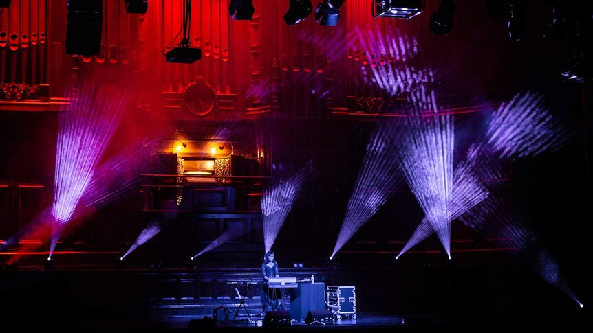 Stage in darkened town hall, with young woman DJing on stage lit by red and purple lights, and a massive organ behind her.