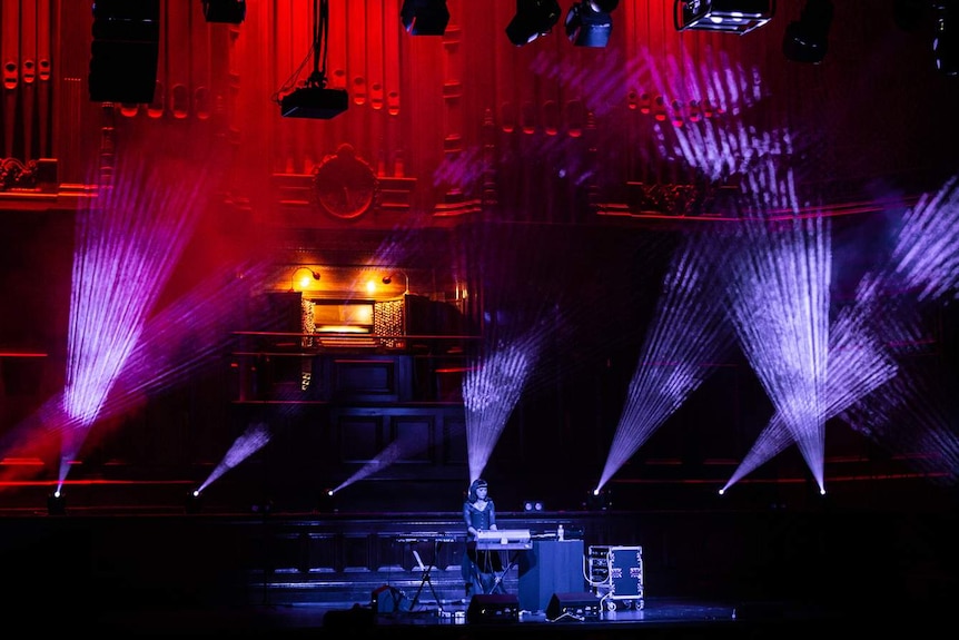 Stage in darkened town hall, with young woman DJing on stage lit by red and purple lights, and a massive organ behind her.