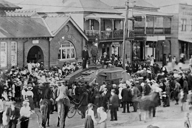 A black-and-white photo shows a group of people gathered around a building in an area town in 1918.