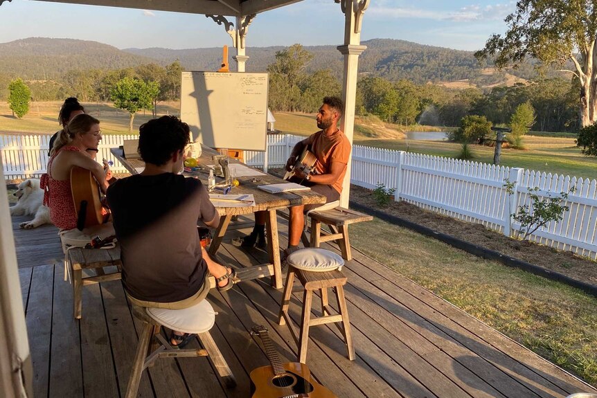 Artists Marcus Corowa, Irena Lysiuk and Jonathan Hickey sit together on a porch practising music while holding instruments