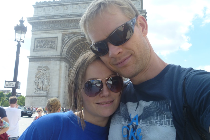 A couple are smiling and standing in front the large Arc de Triomphe monument in Paris.