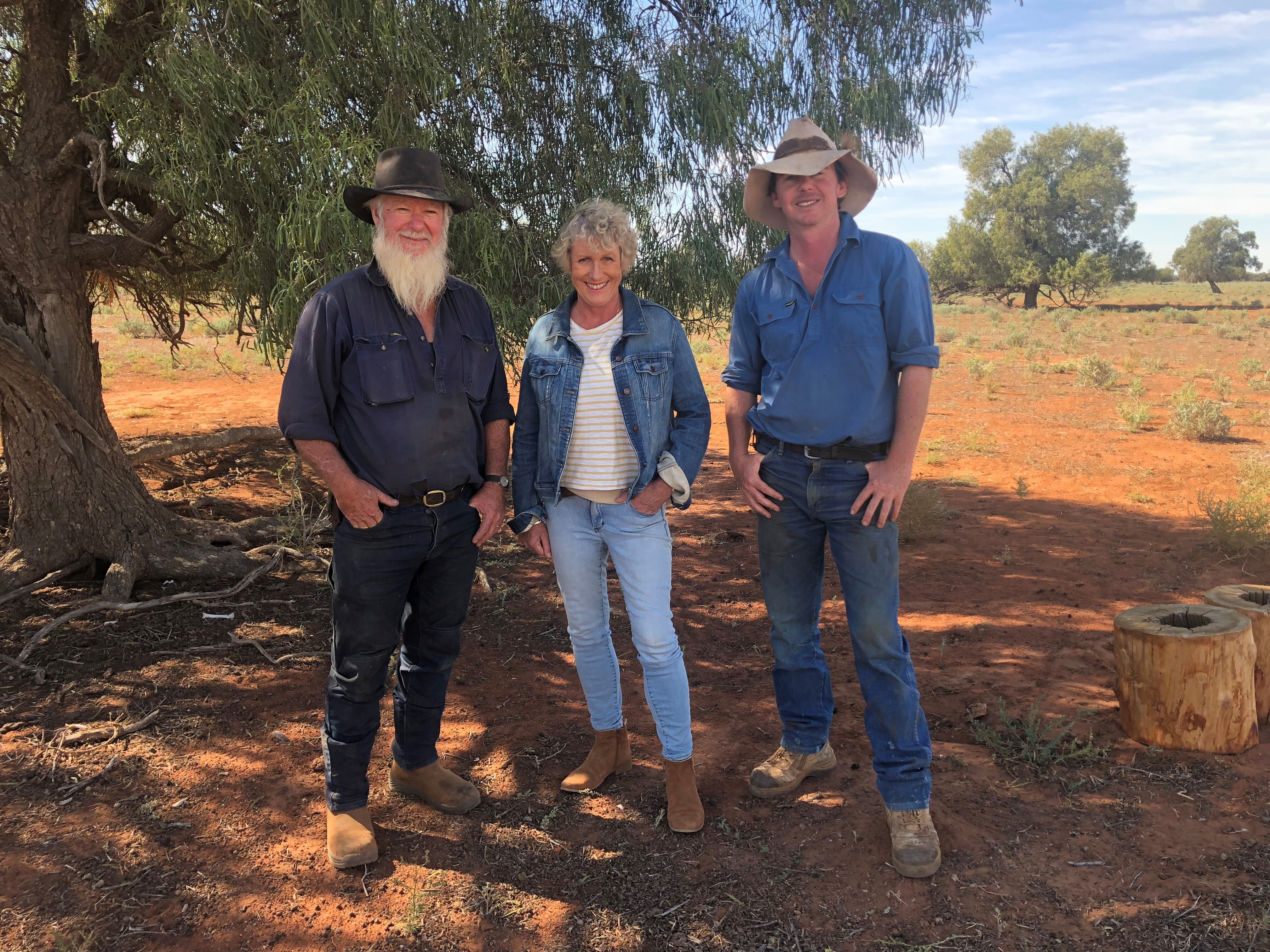 Two men and one woman standing in a red dirt hay field underneath a tree, smiling. 