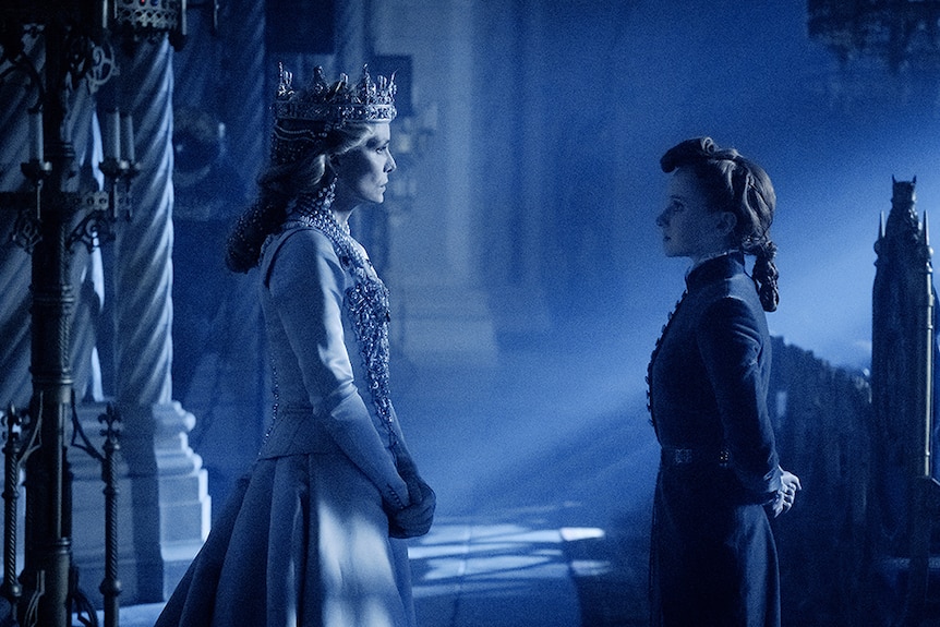Michelle Pfeiffer wears crown and ornate jewelled gown and stands facing Jenn Murray in dimly lit medieval or gothic style room.