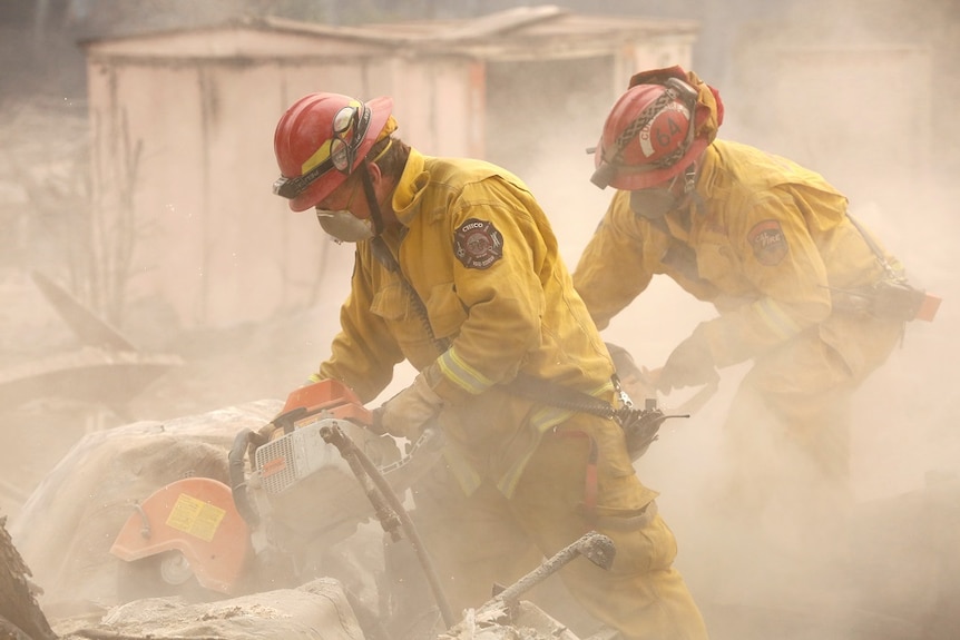 Two firefighters in full gear use a demolition saw to cut through the charred wreckage of a home.