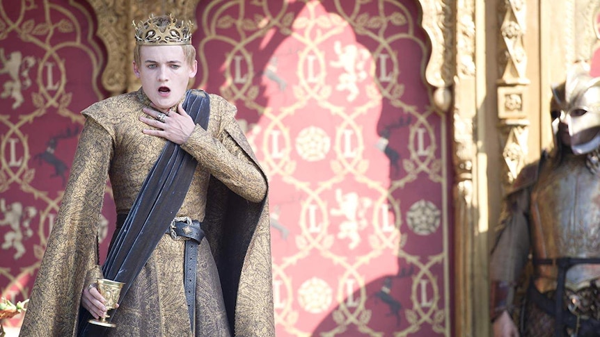 Joffrey Lannister clutches his throat as he wears golden robes and holds a poisoned goblet in his other hand.