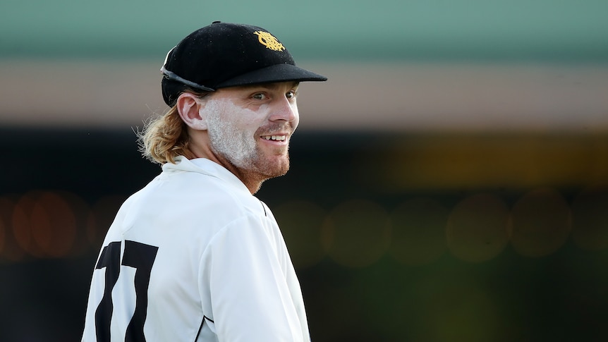 A man with long hair and a cricket cap smiles during a game. 