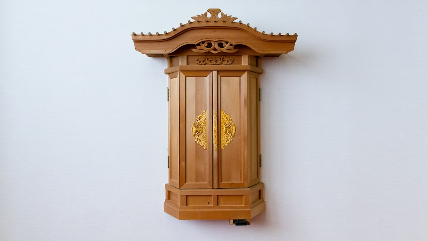 A small brown, wooden cabinet with slim double doors on the front hangs on a pale wall.