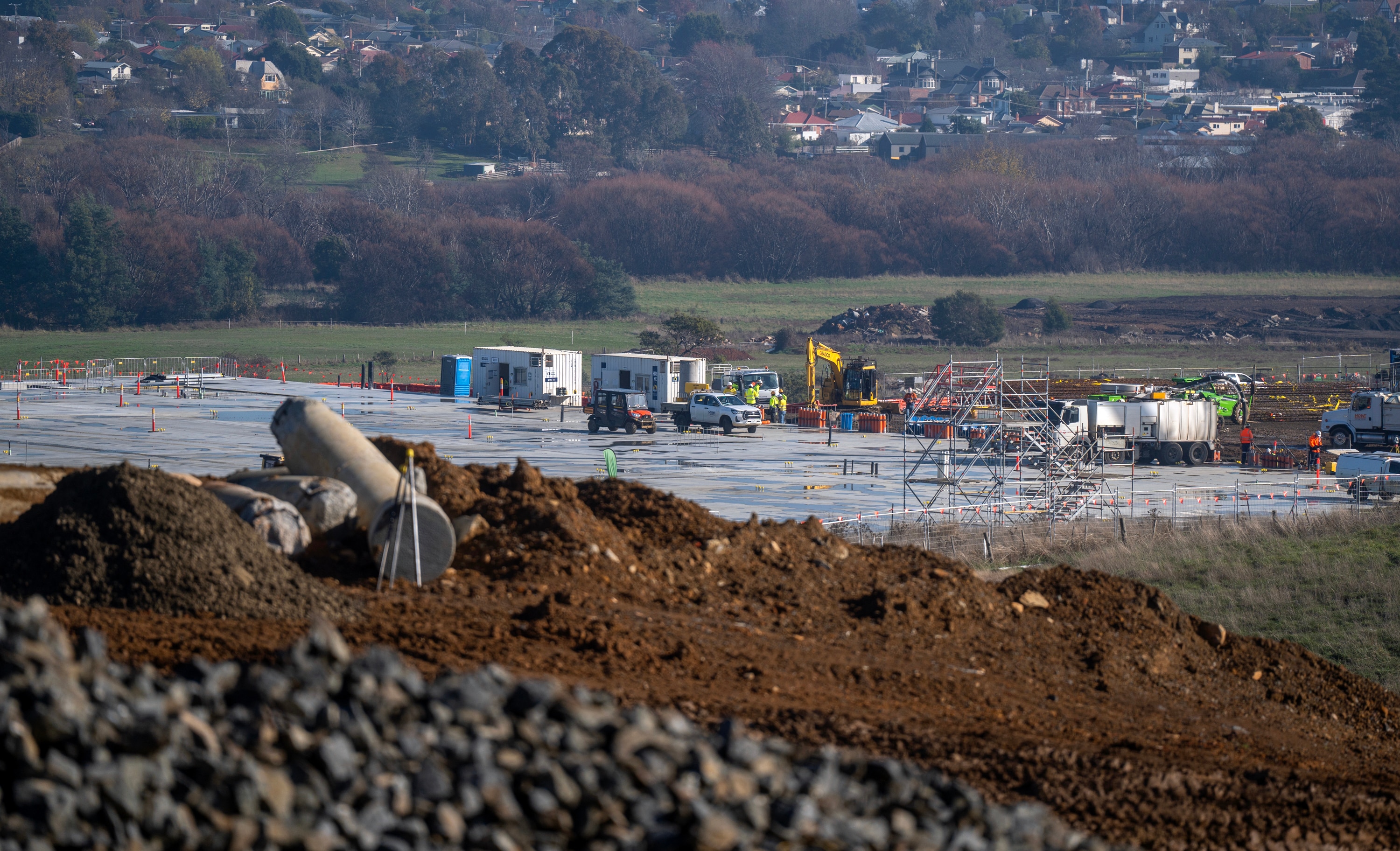 Piles of rocky dirt foregounds a construction site with yellow machinery in a valley dotted with homes.