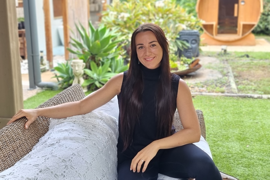 A young woman with long dark brown hair and a black shirt smiling and sitting in her backyard