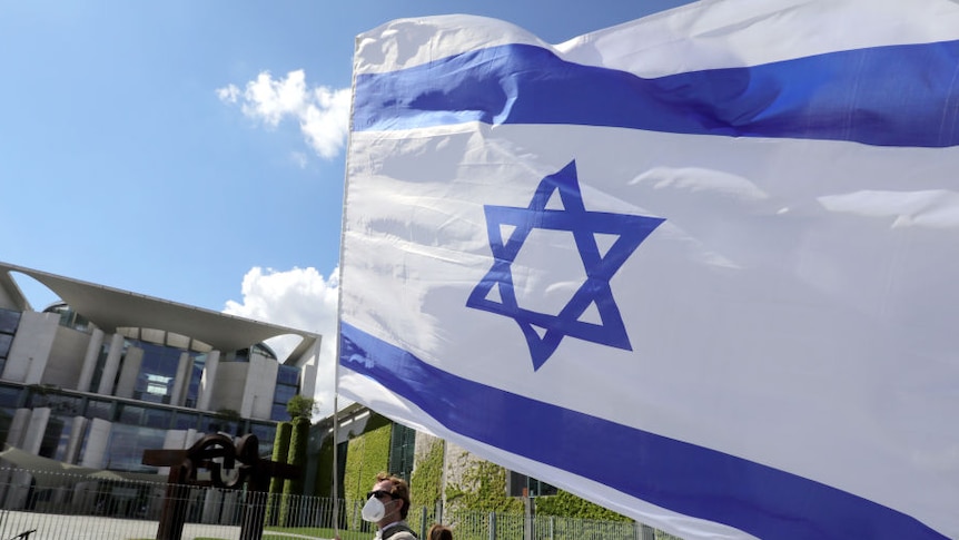 Man carries large Israeli flag flying in the wind in front of fancy building