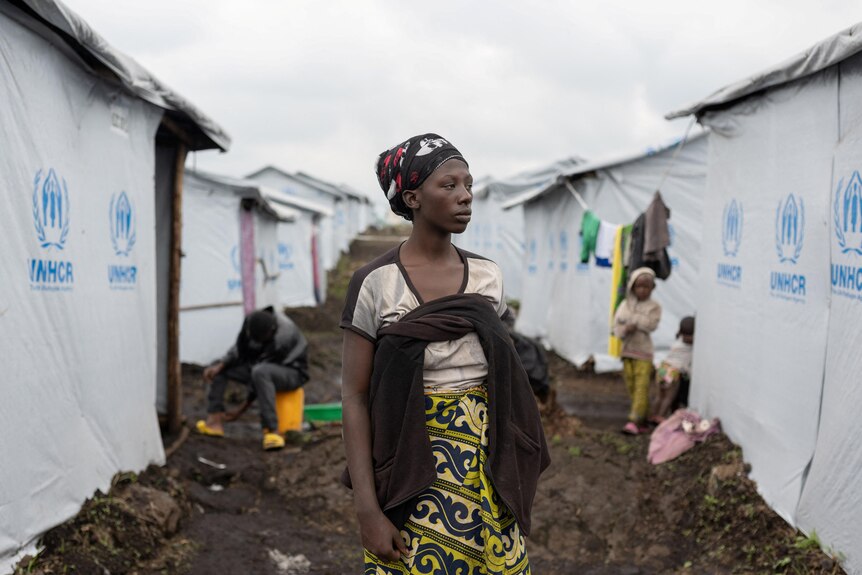 A displaced woman stands in front of a row of tents in a muddy area.