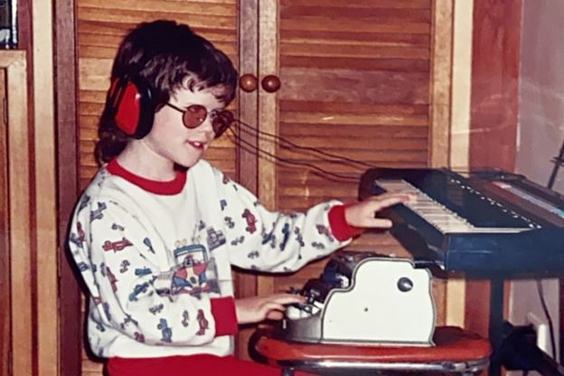 A little boy wearing glasses and over-ear headphones sits at a small wooden stool playing the keyboard.