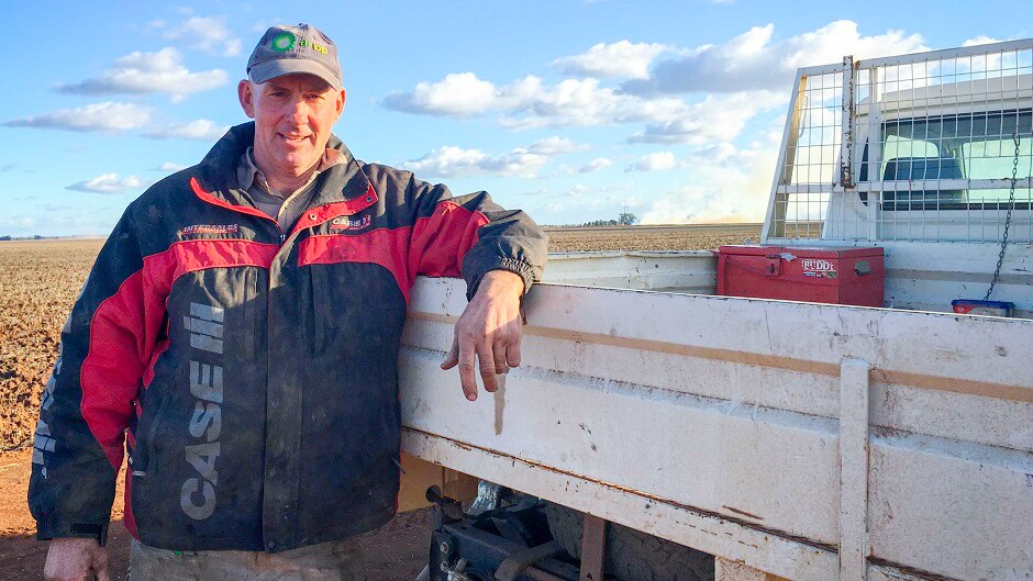 Man in blue hat leans on tray of his ute, next to a bare paddock.