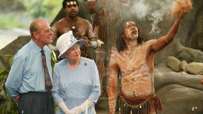 Two people in formal clothes watch an Indigenous man in body paint and tribal dress using smoke