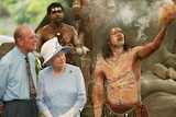 Two people in formal clothes watch an Indigenous man in body paint and tribal dress using smoke