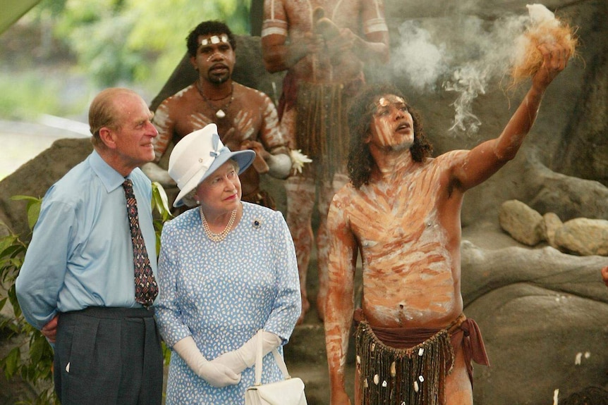 Two people in formal wear watch an Aboriginal man in body paint and tribal dress using smoke.