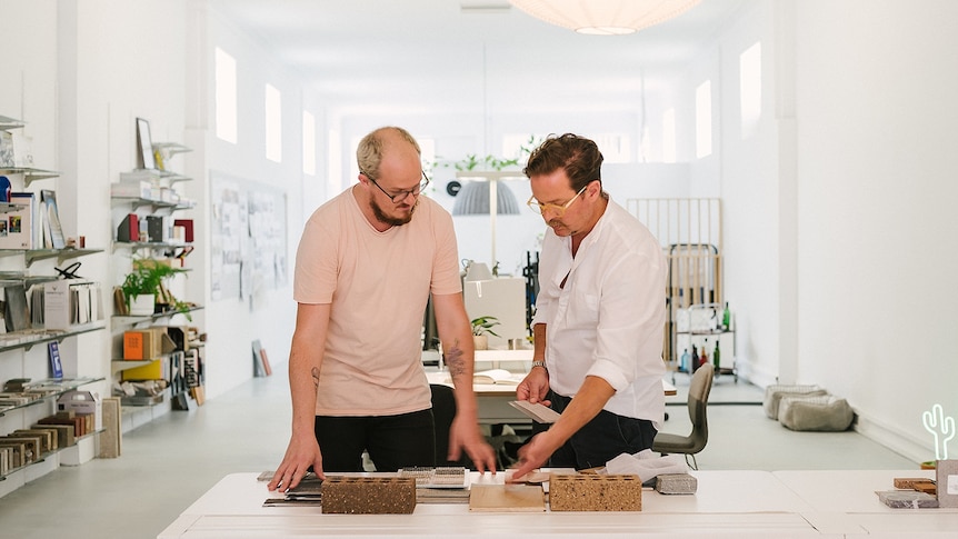 You see two men in light pastel colours looking over a desk with architectural models in a bright, white office.