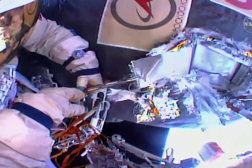 One cosmonaut holds back the heat shield while the other uses a snipping tool.