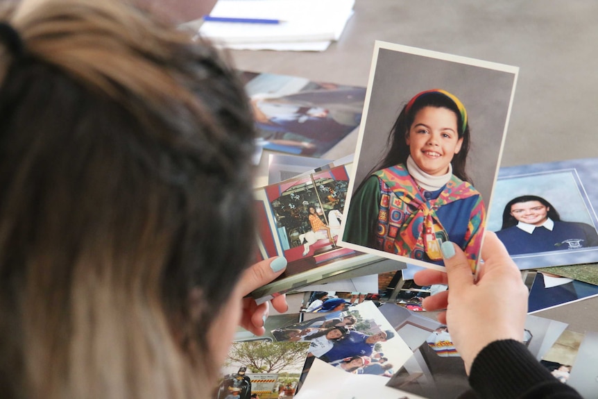 A woman holds photos of herself as a young girl while looking through photos sprawled across a table.