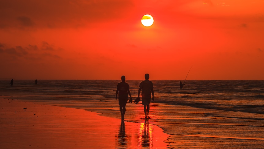 A silhouette of a couple walking on a beach, everything is red at sunset, people fishing in the background