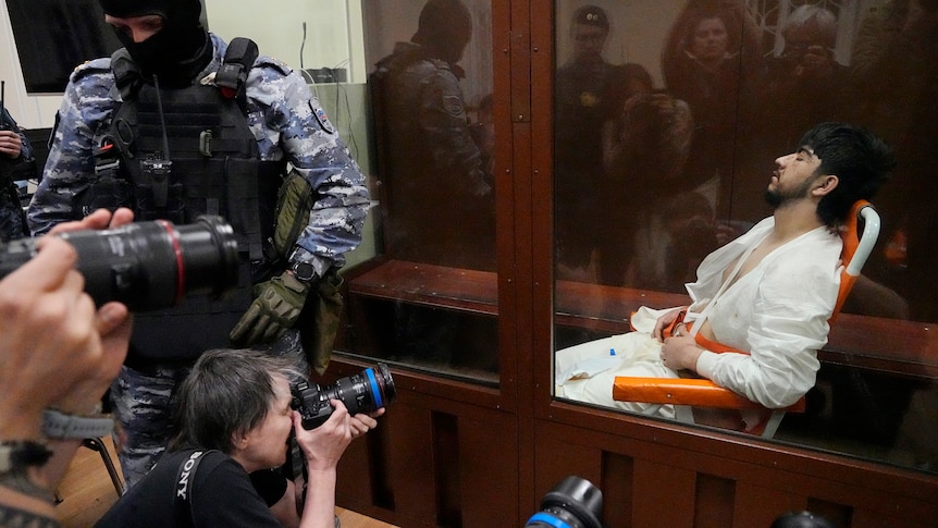 A man in a white jumpsuit reclines in a chair within a glass cage as photographers gather outside.