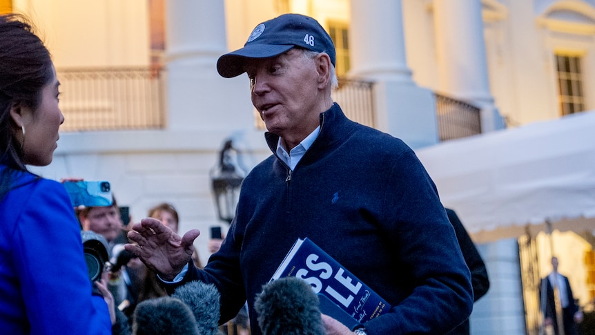 A medium shot of Joe Biden wearing a hat and speaking with members of the media outside the White House.