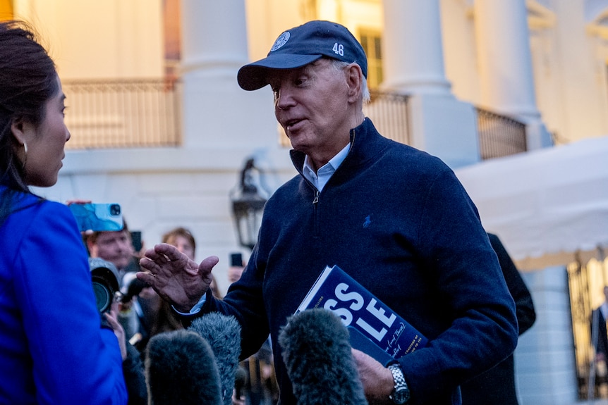 A medium shot of Joe Biden wearing a hat and speaking with members of the media outside the White House.
