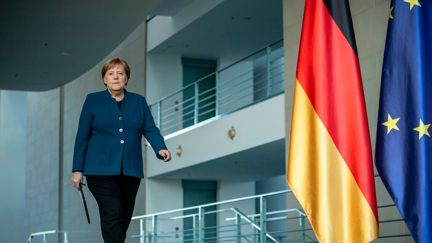 Angela Merkel walks in a navy blazer down a corridor with a large German and EU flag.