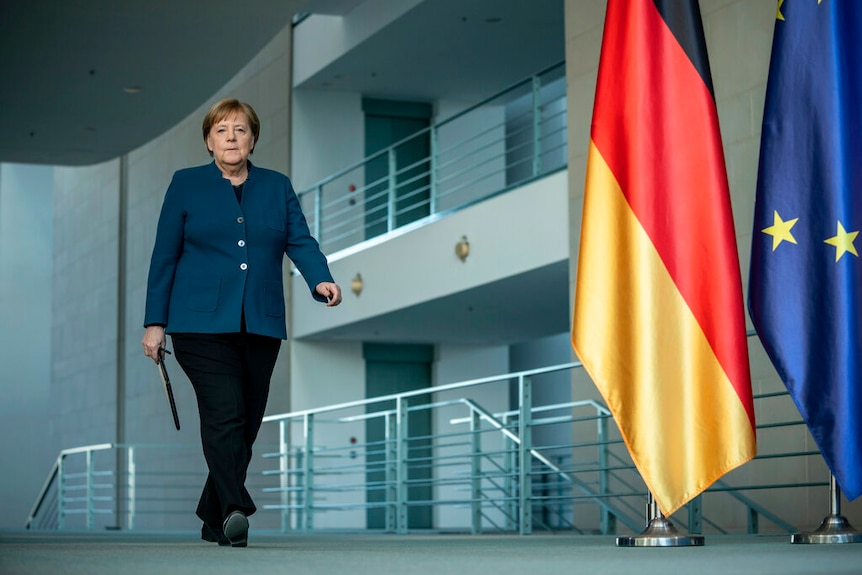 Angela Merkel walks in a navy blazer down a corridor with a large German and EU flag.