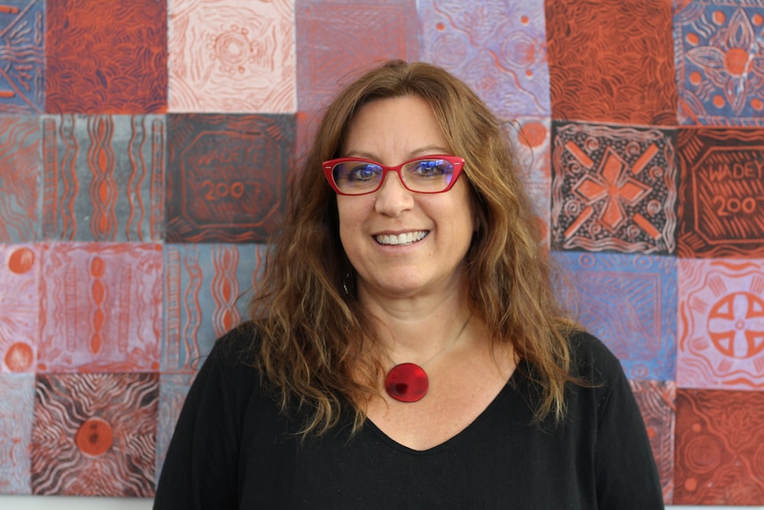 headshot of smiling woman with red glasses, shoulder length light brown hair in from of red, purple square pattern painting