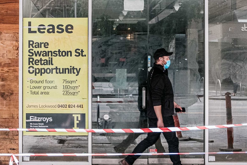 A man in a mask walks past an empty shop with a for lease sign on it.