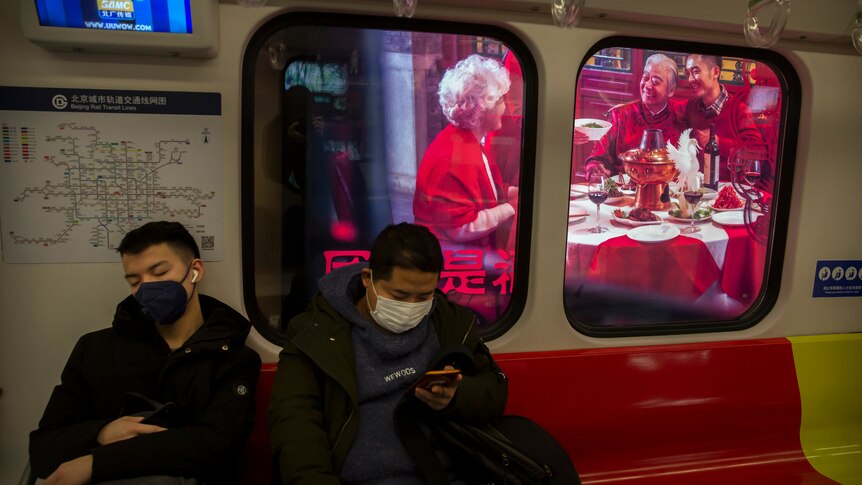 Two men wear masks on a subway train as it stops near a billboard showing a family having a New Year's banquet meal.