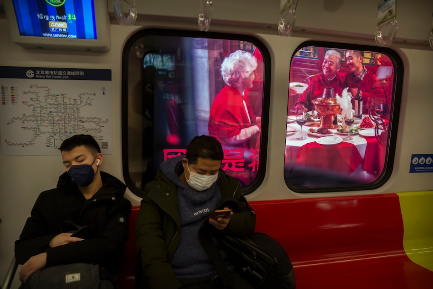 Two men wear masks on a subway train as it stops near a billboard showing a family having a New Year's banquet meal.