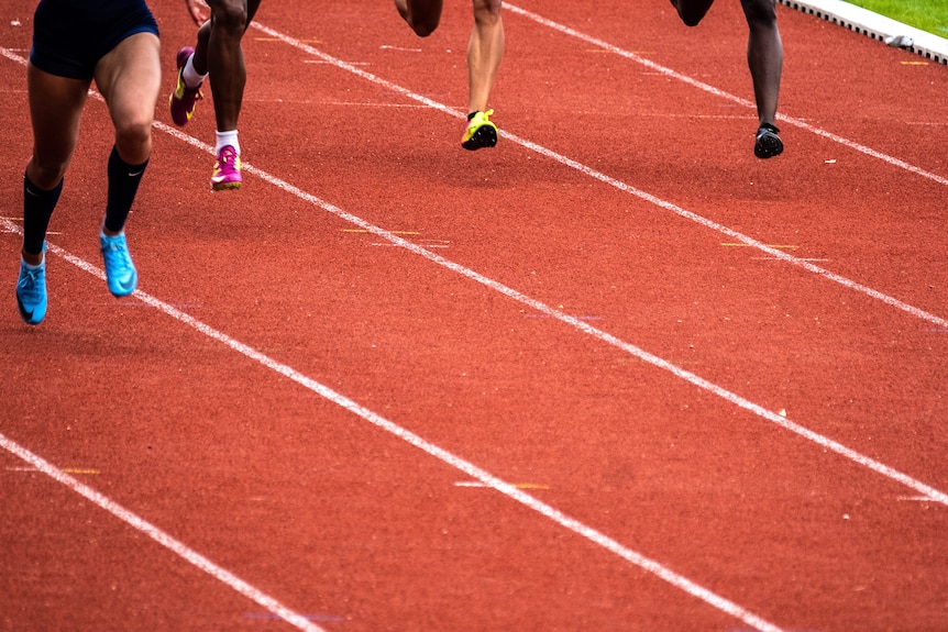 Four people's legs can be seen running down a red running track with lane markings. They are wearing running shoes.