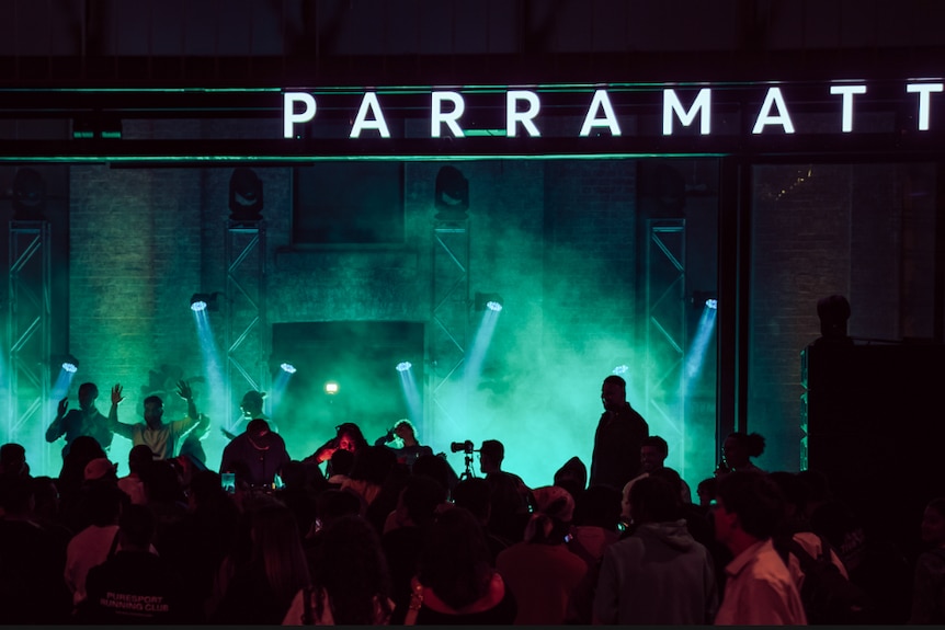 A silhouette of a crowd in front of a greenlit dancefloor. A lit up sign reads ‘Parramatta’. 