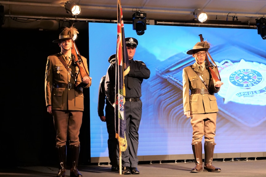 One man in police uniform holds the police flag in front of him, flanked by two ADF personnel in uniform with rifles on shoulder