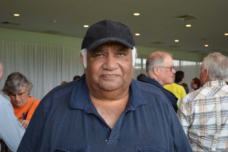 An Aboriginal man in a navy polo shirt and black cap looks seriously at the camera.