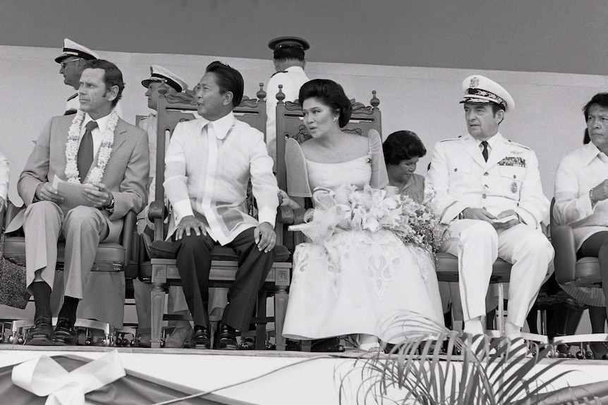 A black and white photo of Ferdinand and Imelda Marcos sitting on a stage, surrounded by men in military uniform