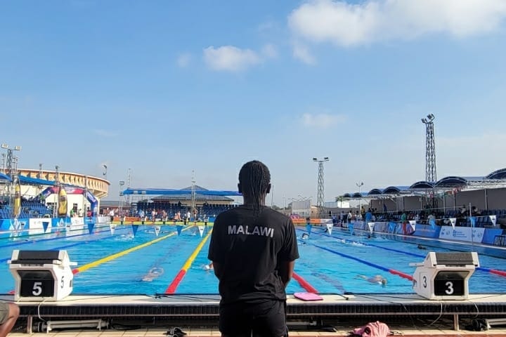 A woman in a black t-shirt with Malawi on the back of it looks out at a pool.