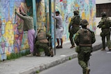 Soldiers in green camouflage uniforms and holding rifles frisk two men up against a colourful brick wall.