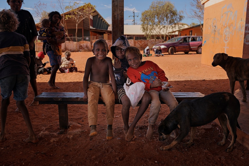 Three kids sitting on a wooden bench smile at the camera in the remote community of Kintore.