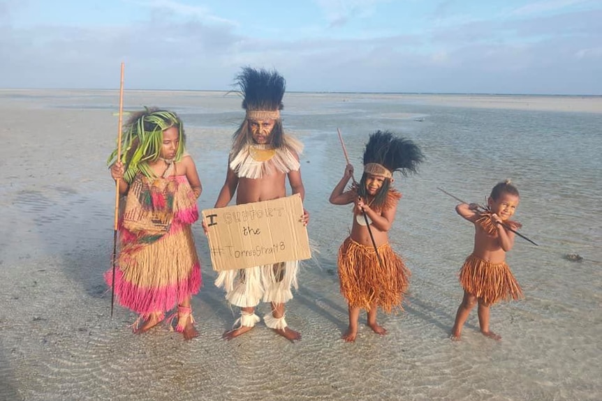 Four young children in traditional Torres Strait Islander dress stand on beach holding sign calling for climate action.
