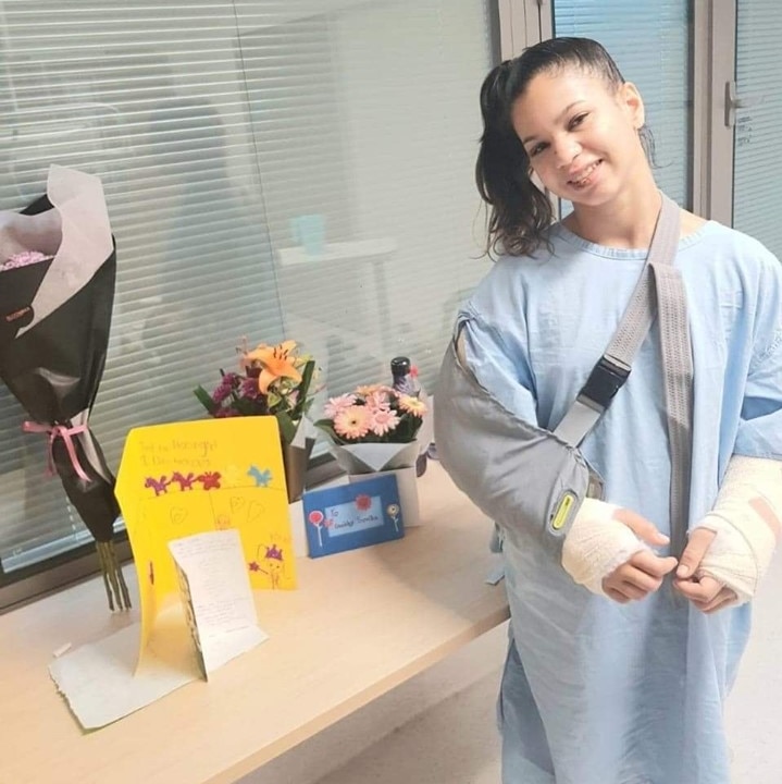 Gabby Souza stands up wearing a hospital gown with both wrists covered in casts